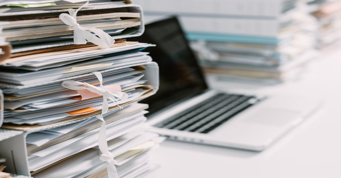 Stacks of paperwork tied down with strings sitting on a white desk. There is an open laptop next to the documents.