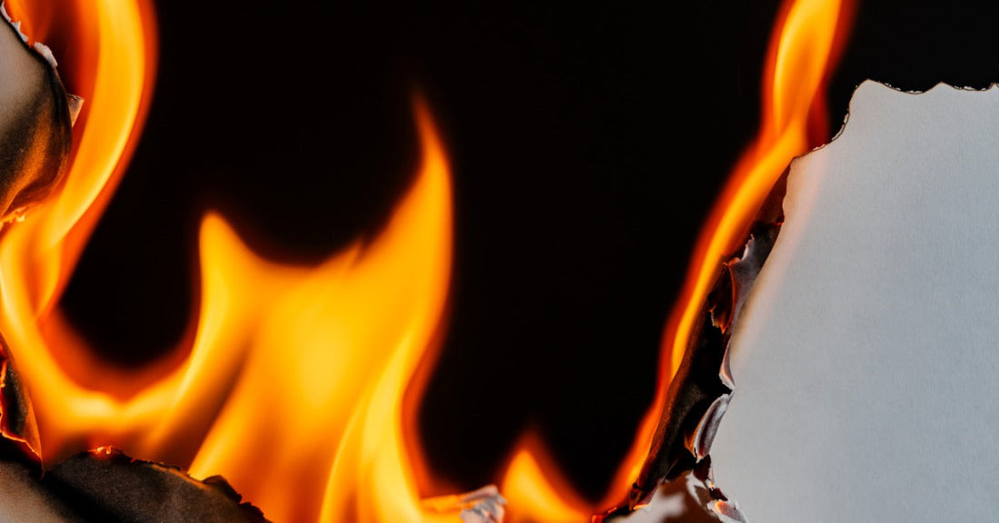 A sheet of white paper has black edges as a bright orange and red flames burns the sheet against a black background.
