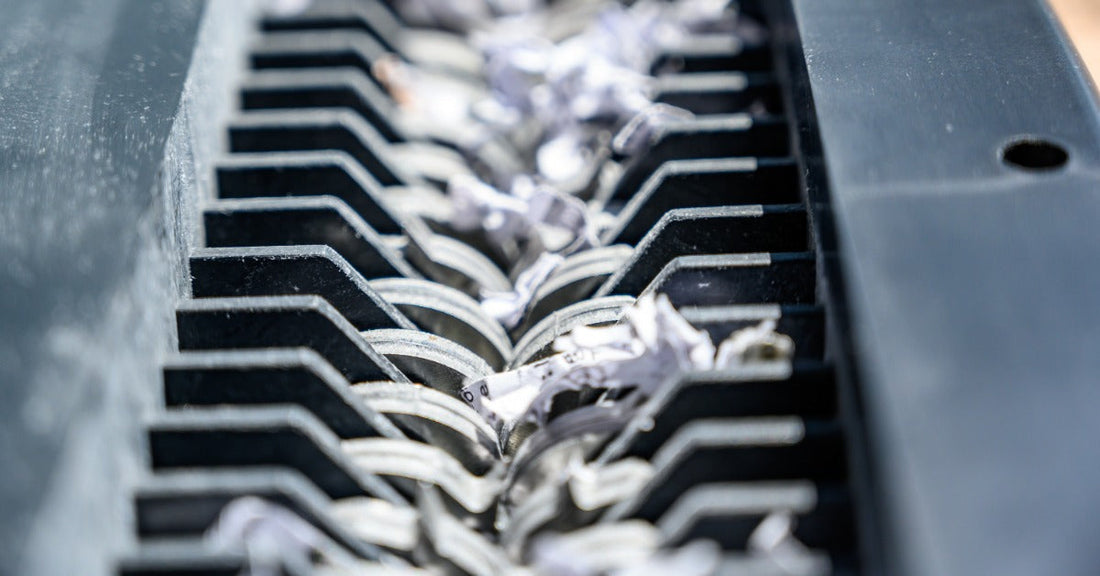 A close-up of the cutting blades of a paper shredder. Small pieces of white paper are moving through the blades.