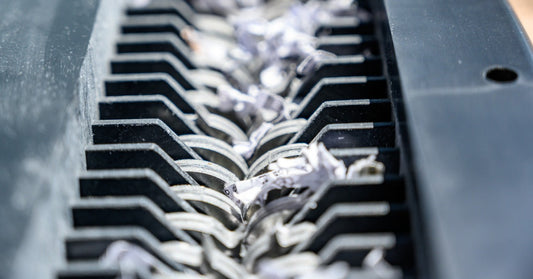 A close-up of the cutting blades of a paper shredder. Small pieces of white paper are moving through the blades.