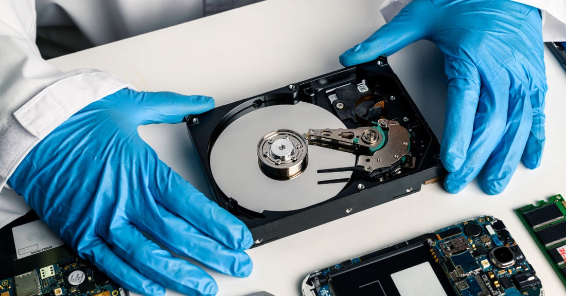 Two blue gloved hands are hovering beside an open hard drive. Disassembled computer parts are scattered on the table.