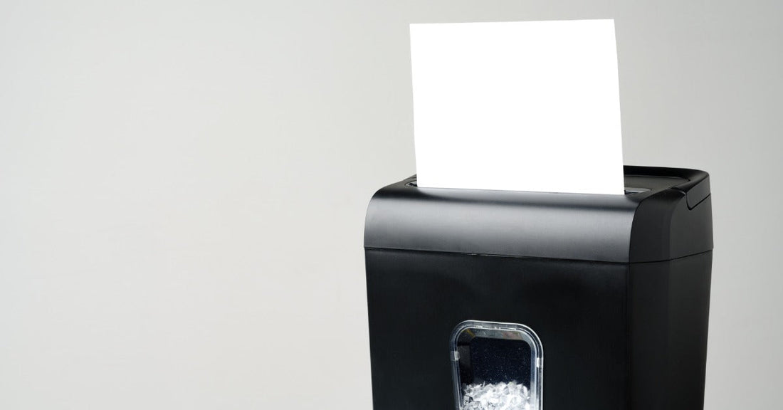 A black paper shredder has a white sheet inserted into the top slot against a plain gray background.