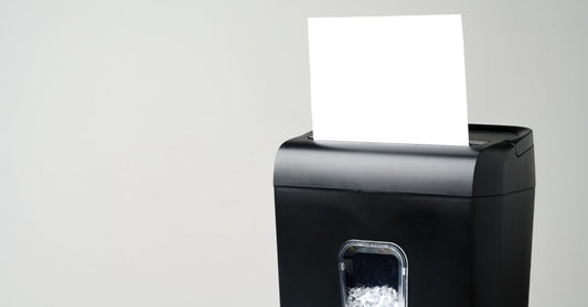 A black paper shredder has a white sheet inserted into the top slot against a plain gray background.