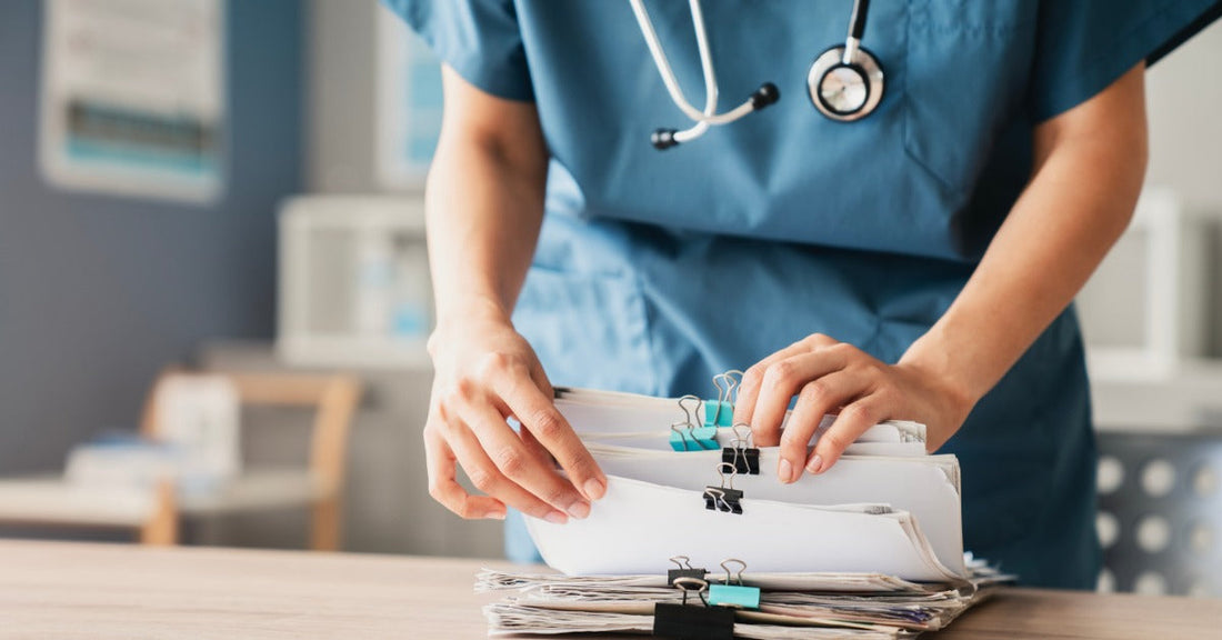 A healthcare professional wearing blue scrubs picks through stacks of documents secured by binder clips.