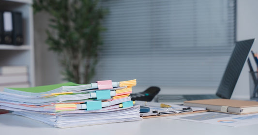 A stack of papers with colorful sticky tabs and binder clips on an office desk. A clipboard and laptop are in the background.