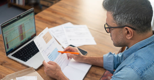 A man wearing glasses is holding an orange pen and a utility bill. A laptop is open and displays a data sheet.