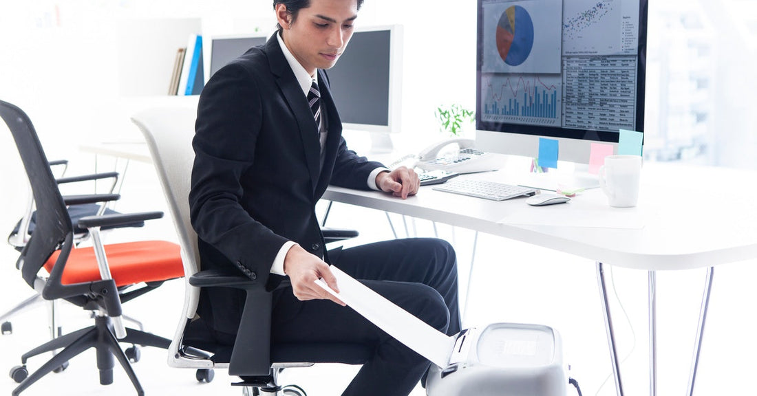 An office worker sitting at his desk with a chart on his monitor. He is shredding a sheet of paper underneath his desk.