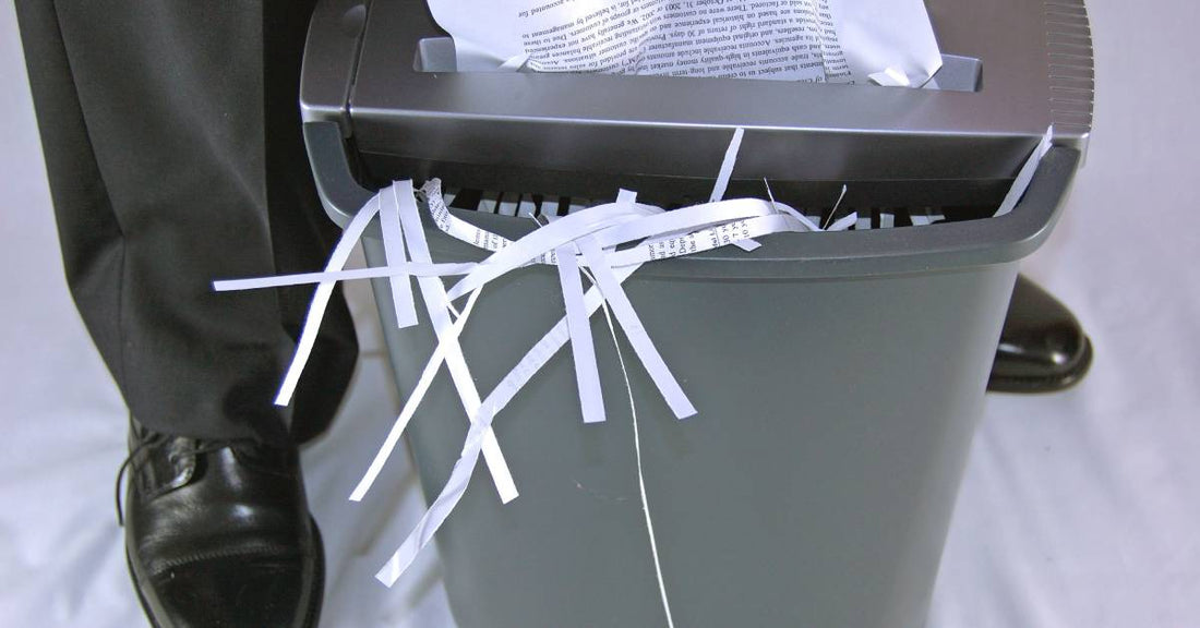A crumpled paper inserted into a paper shredder next to a person. There are strips of paper sticking out from the bin.