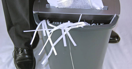 A crumpled paper inserted into a paper shredder next to a person. There are strips of paper sticking out from the bin.
