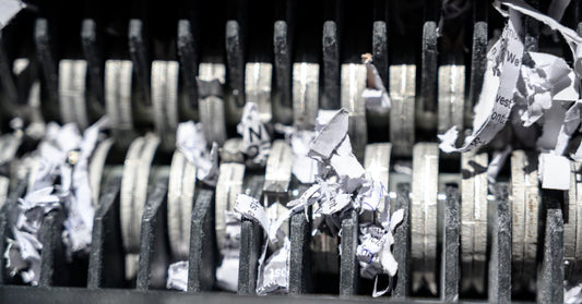 The silver, metal teeth of a paper shredder at close range. Tiny pieces of torn paper are lodged in the machine.