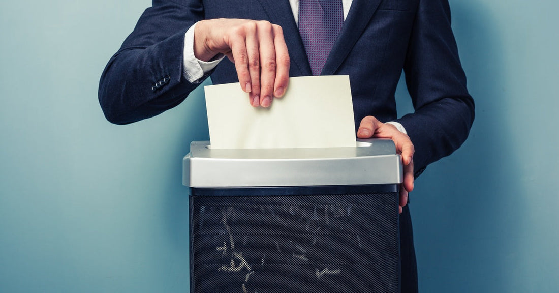A person wearing a suit places a sheet of paper into a paper shredder. Small pieces are in the shredder reservoir.