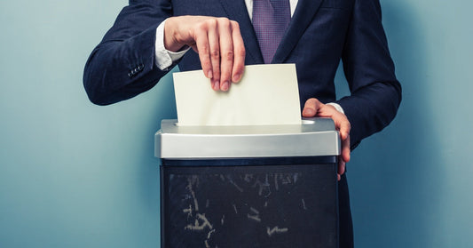 A person wearing a suit places a sheet of paper into a paper shredder. Small pieces are in the shredder reservoir.