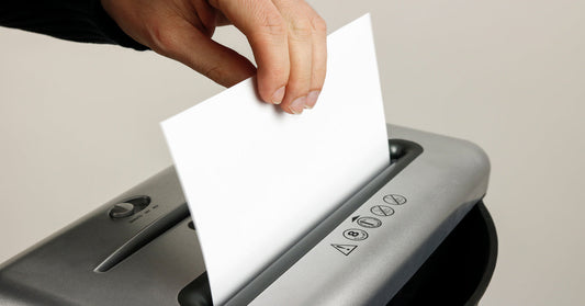 A close-up of a person inserting one sheet of paper into a silver paper shredder. The background is beige.