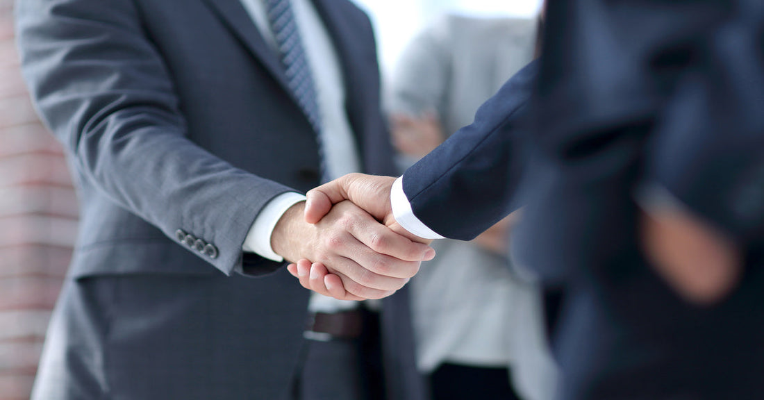 A close-up of two men shaking hands. One person wears a gray suit with a tie, and the other wears a navy suit.