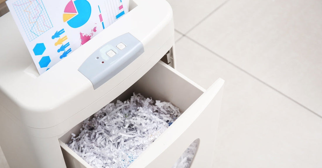 A white paper shredder shreds a sheet with various color-coded graphs. The discard bin is open and full of strips of paper.