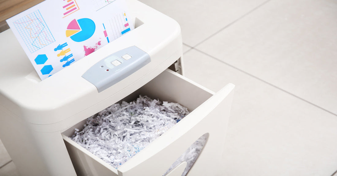 A paper with multicolored graphs is in the feeder of a paper shredder. The bin of the machine is open and full of strips.