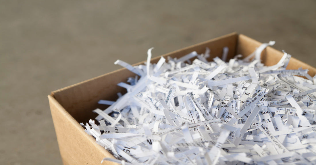 A cardboard box is overflowing with strip-cut paper shreds. Unreadable black text is on the sheets of paper.
