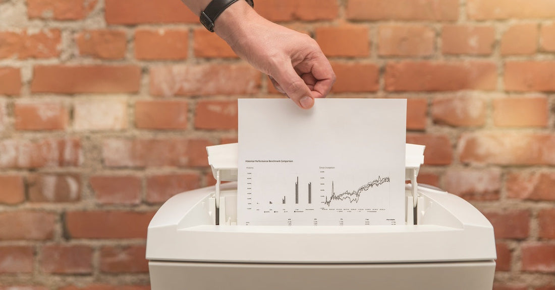 A person wearing a watch inserts a paper with graphs into a small paper shredder. A brick wall fills the background.