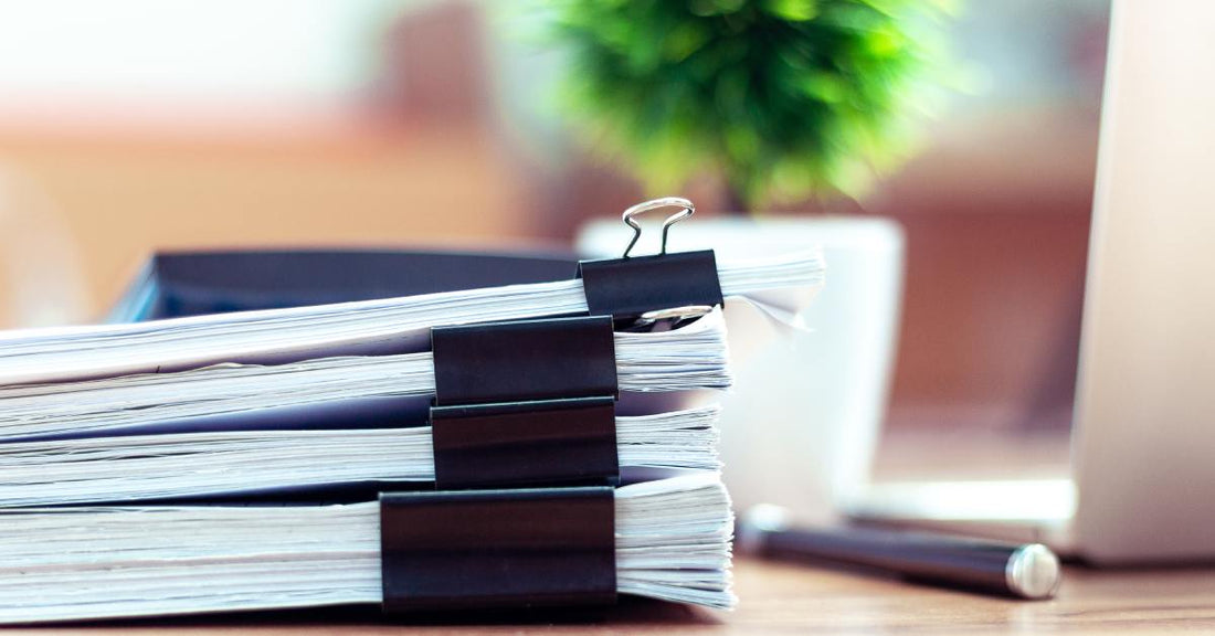 Four piles of paper secured with black binder clips are stacked on top of each other on a wooden desk.