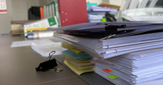 A tall stack of papers and folders are lying on a desk. Binders, notecards, and other office supplies are in the background.