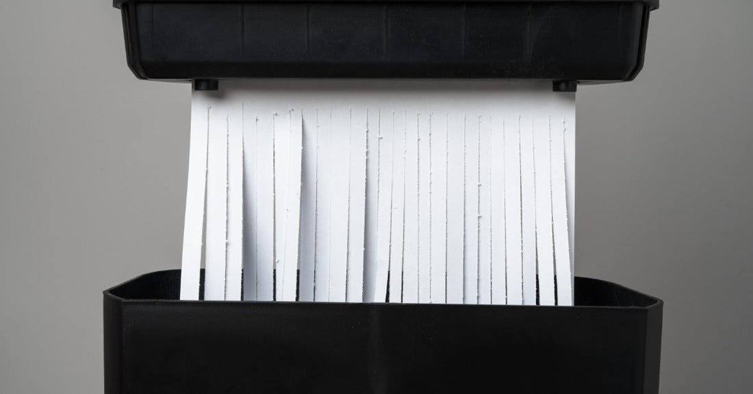 White sheets of paper sliced into strip-cut shreds are existing a black shredder that's against a gray background.