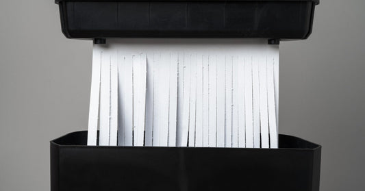 White sheets of paper sliced into strip-cut shreds are existing a black shredder that's against a gray background.