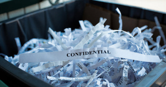 A close-up of strip-cut paper shreds in a black bin. One long, thick strip of paper reads, "CONFIDENTIAL."
