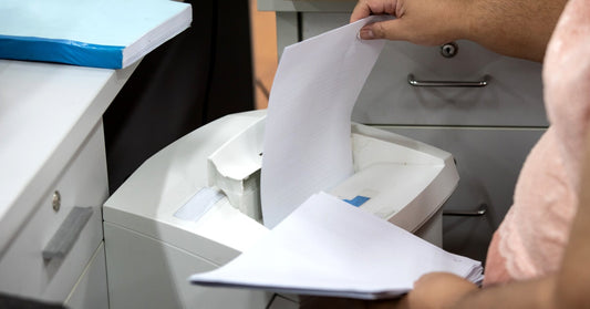 A person feeds a sheet of paper into a paper shredder. File cabinets and notebooks surround the shredder.