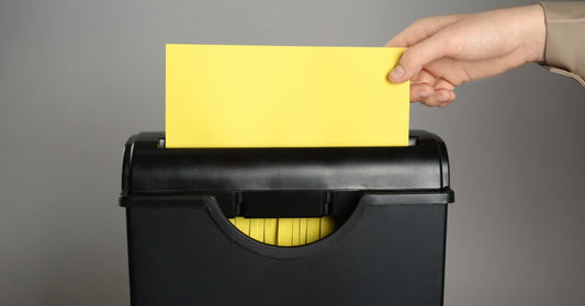 A hand places a yellow sheet of paper into a small deskside paper shredder against a gray background.