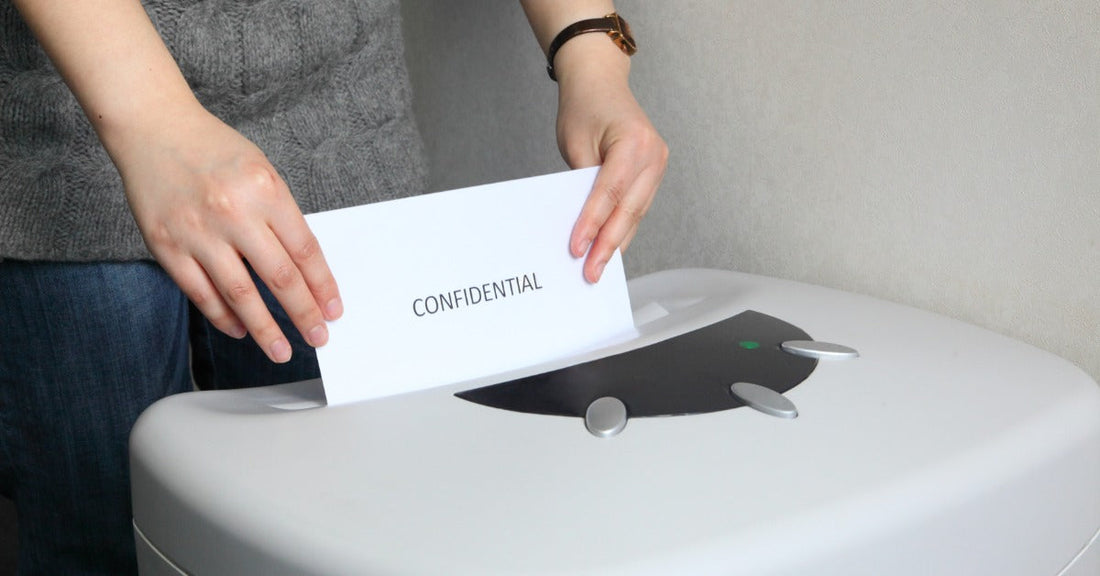A person feeds a white sheet of paper with the text "CONFIDENTIAL" into the opening of a large paper shredder.