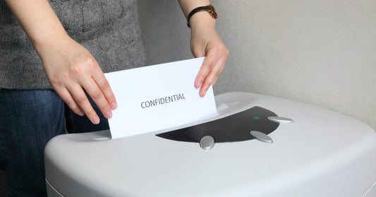 A person feeds a white sheet of paper with the text "CONFIDENTIAL" into the opening of a large paper shredder.