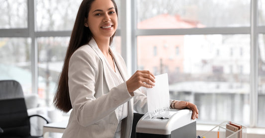  woman wearing a blazer is smiling as she inserts a sheet of paper into a paper shredder on a desk.