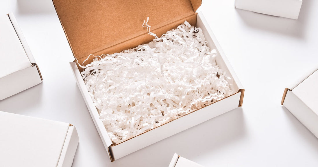 A white cardboard packaging box full of thin strips of shredded paper. Other closed white cardboard boxes sit nearby.