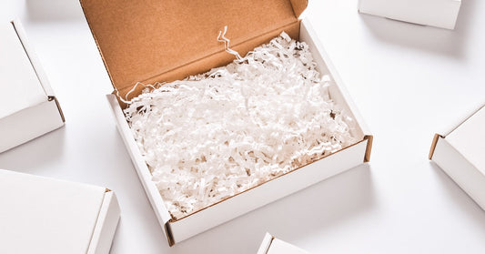 A white cardboard packaging box full of thin strips of shredded paper. Other closed white cardboard boxes sit nearby.