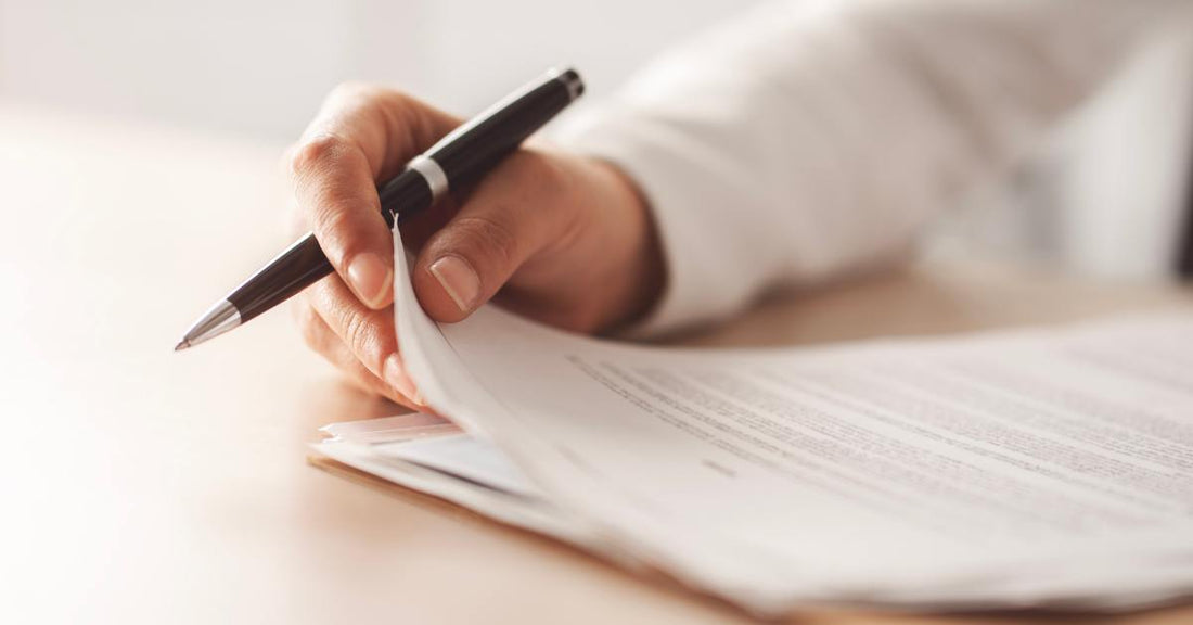 A close-up of a person holding a pen and the corner of a stack of papers. The papers are on a tan desk.